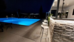 A swimming pool with a stone wall and bar stools on one side, and a patio area with a stone wall and a lamp post on the other.