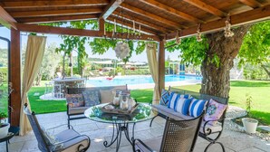 A covered patio area with outdoor furniture, a glass table, and a view of a swimming pool and greenery.