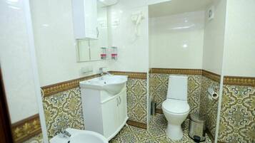 Une salle de bain avec un lavabo, des toilettes et un miroir, tous de couleur blanche. Le sol est recouvert de carreaux à motifs, et il y a une poubelle près des toilettes.