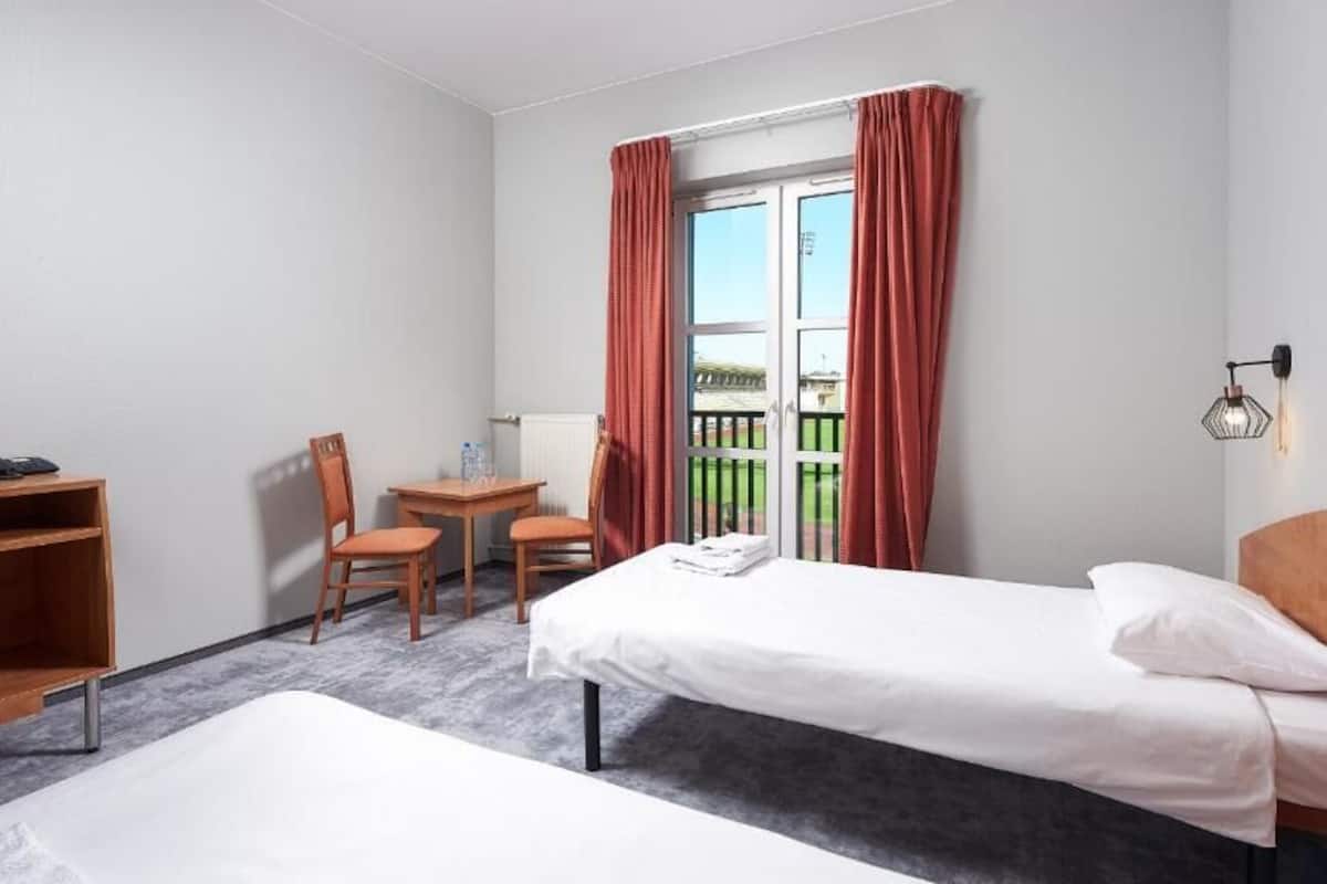 Habitación de hotel con dos camas individuales, un escritorio con silla, una mesita con otra silla, y vista al exterior por una puerta corredera.