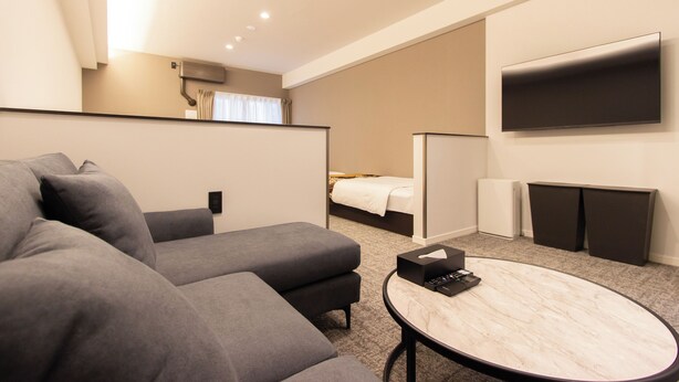 Una habitación de hotel moderna con un sofá gris, una pequeña mesa redonda, una cama con edredón blanco, un televisor montado en la pared y un mueble auxiliar.