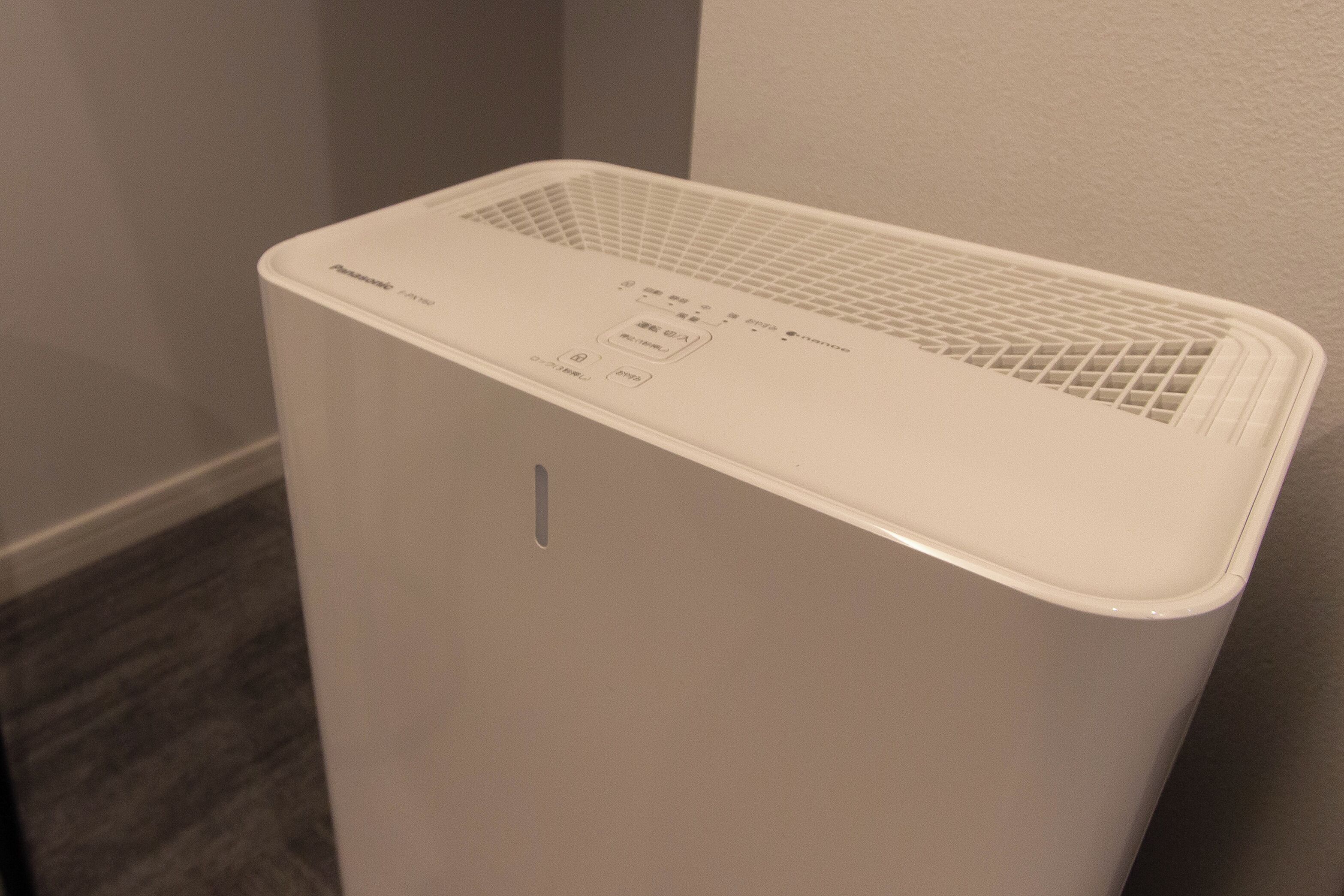 A white portable air purifier with a control panel, placed against a wall in a room with a wooden floor.