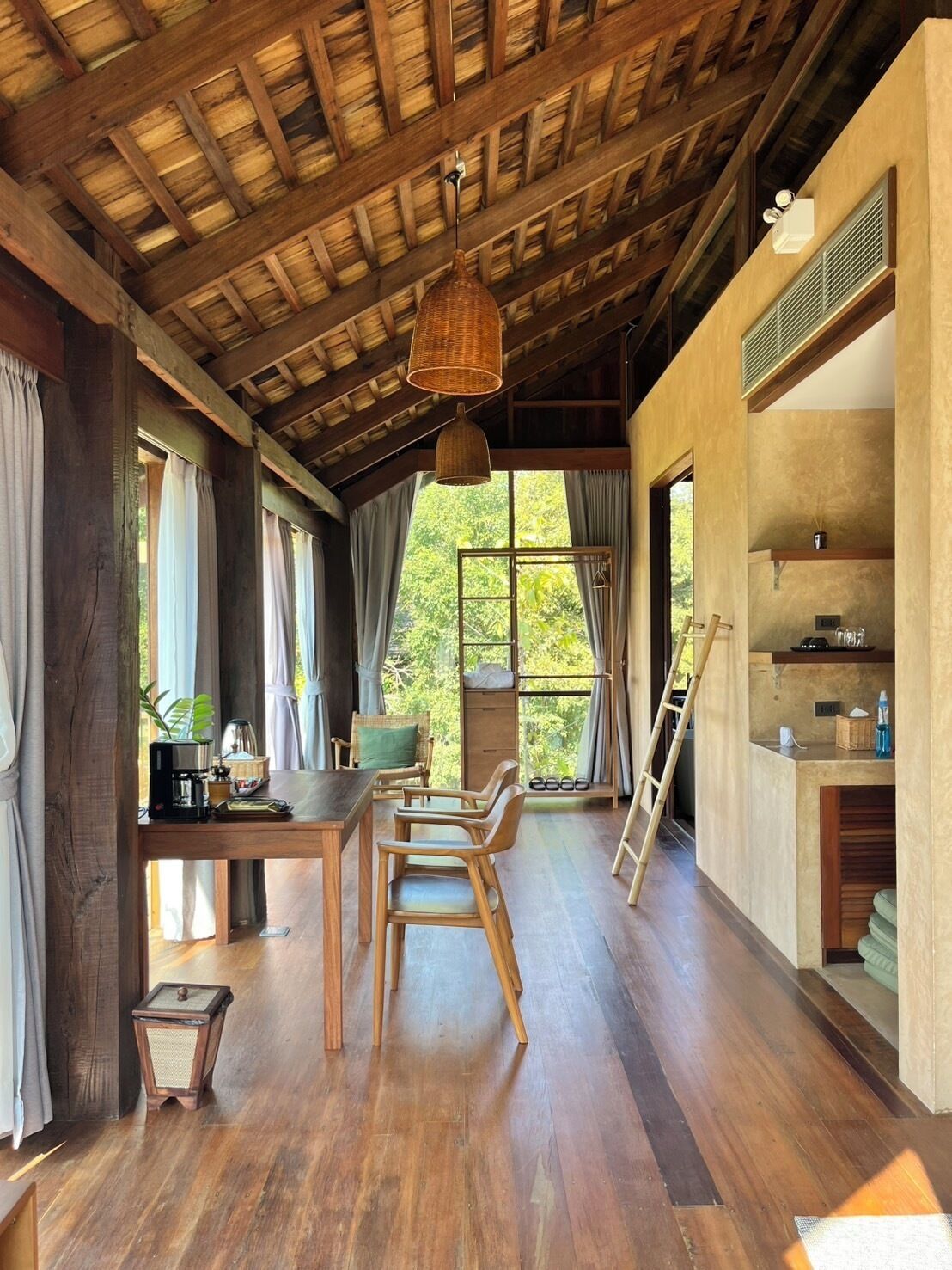 A spacious room with a wooden ceiling, a dining area with a table and chairs, and a large window offering a view of greenery outside.