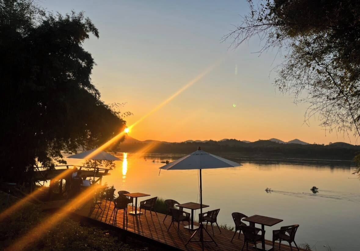 A lakeside dining area with tables and chairs, surrounded by trees, during sunset.
