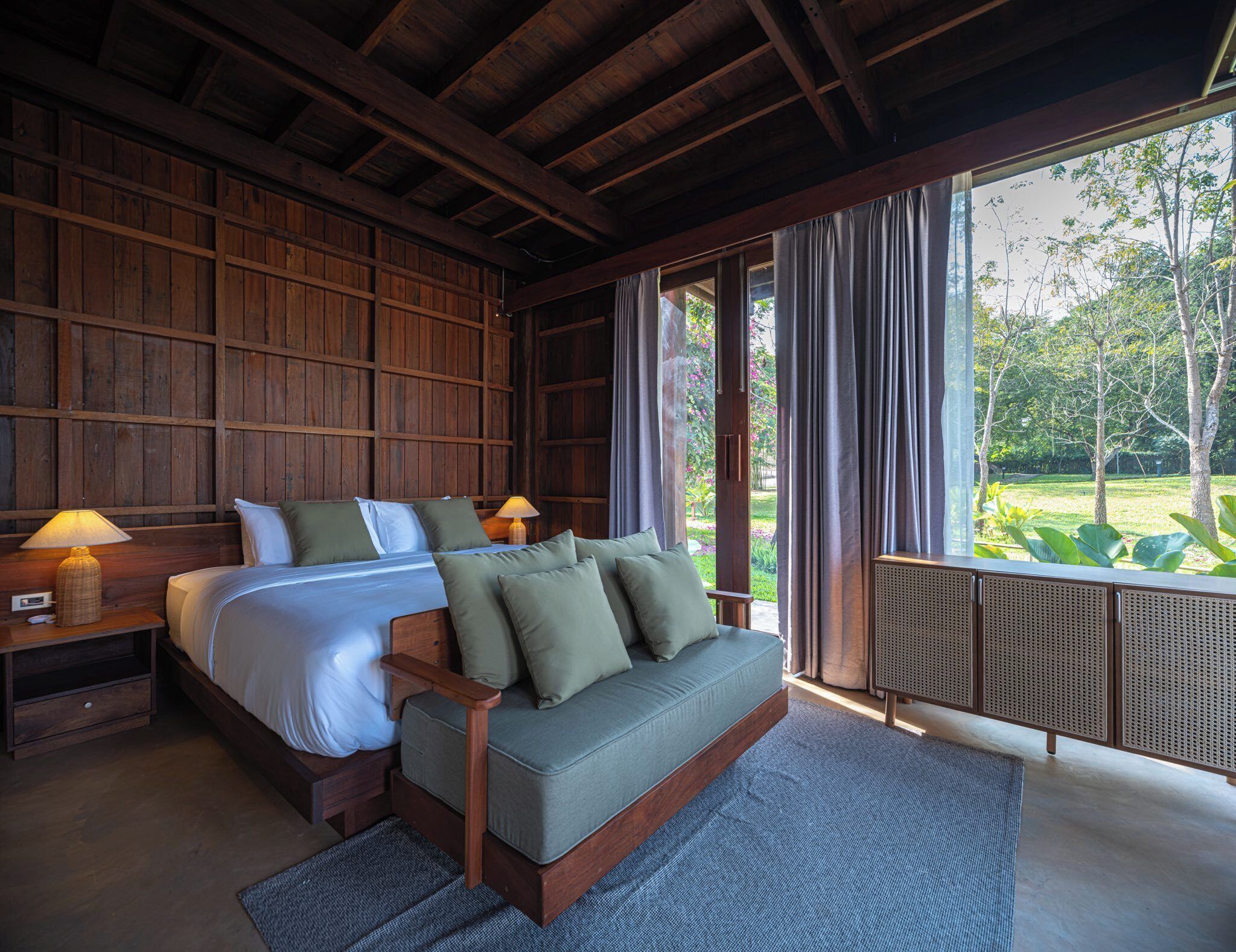A bedroom with a wooden ceiling, a bed with green pillows, a nightstand with a lamp, and a view of a garden through large windows.