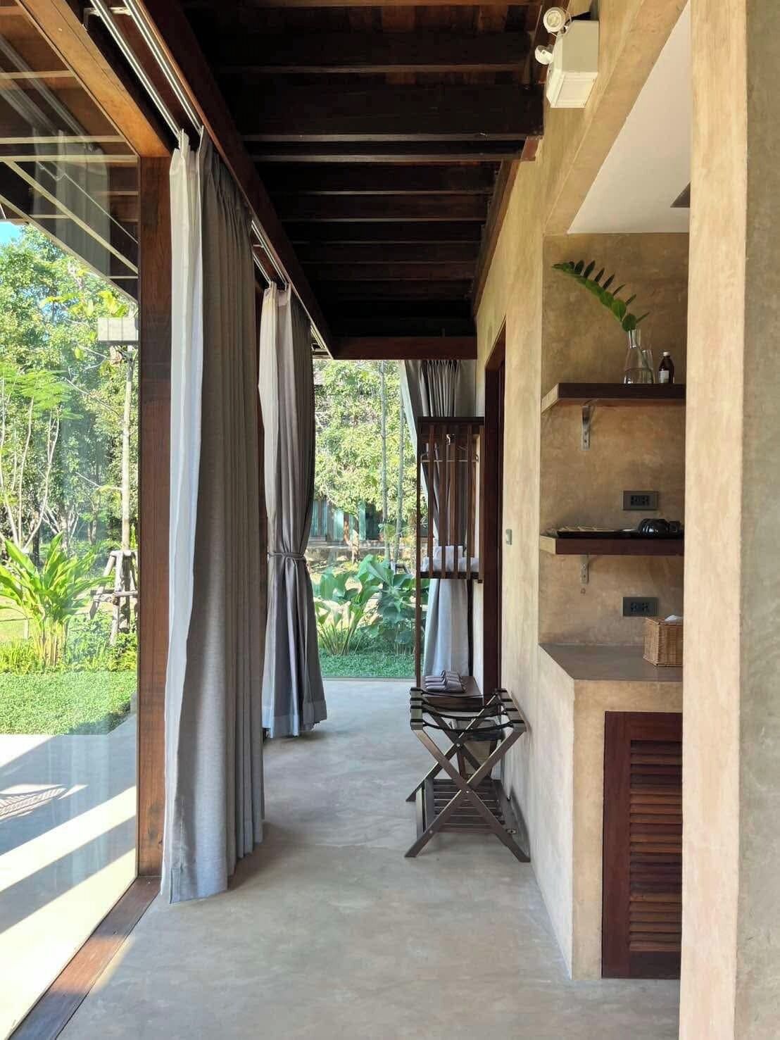 A corridor with a view of a lush garden, a wooden bench, and a shelf with items.