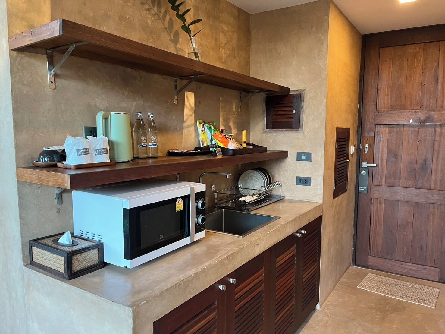 A compact kitchen with a microwave, a coffee machine, and a small sink. There are shelves above the counter holding various items.