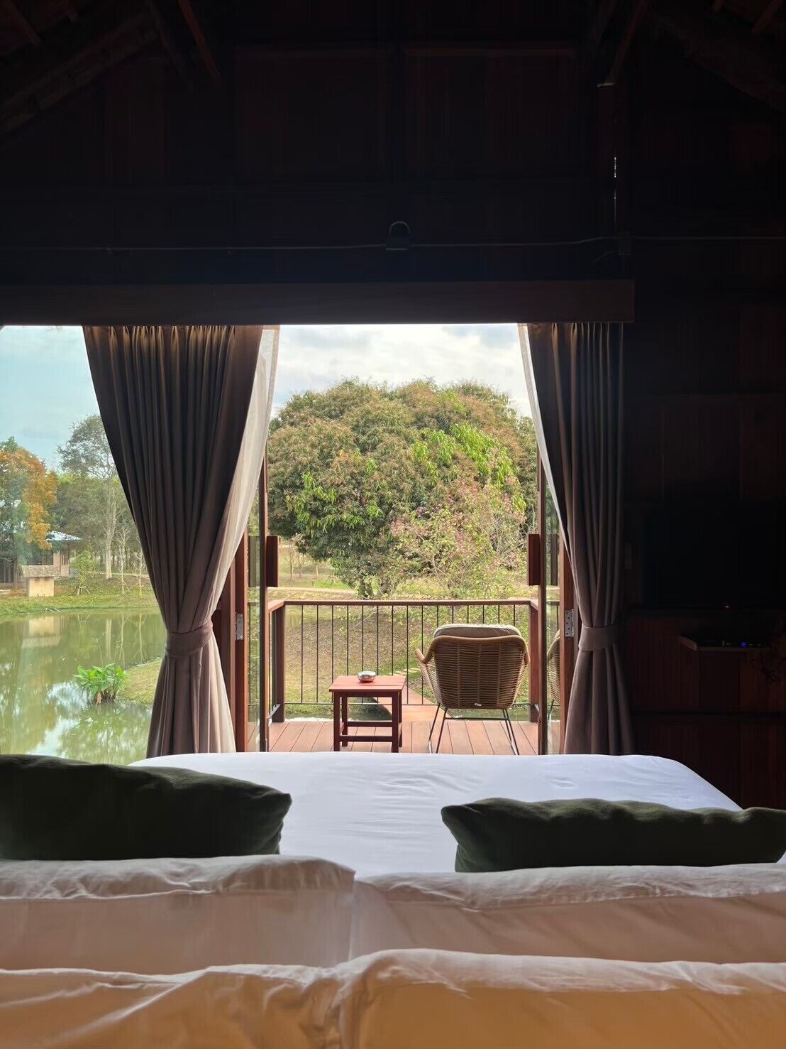 Un dormitorio con una cama grande, dos almohadas, una mesita y una silla. Hay un balcón con vistas a un lago y árboles.