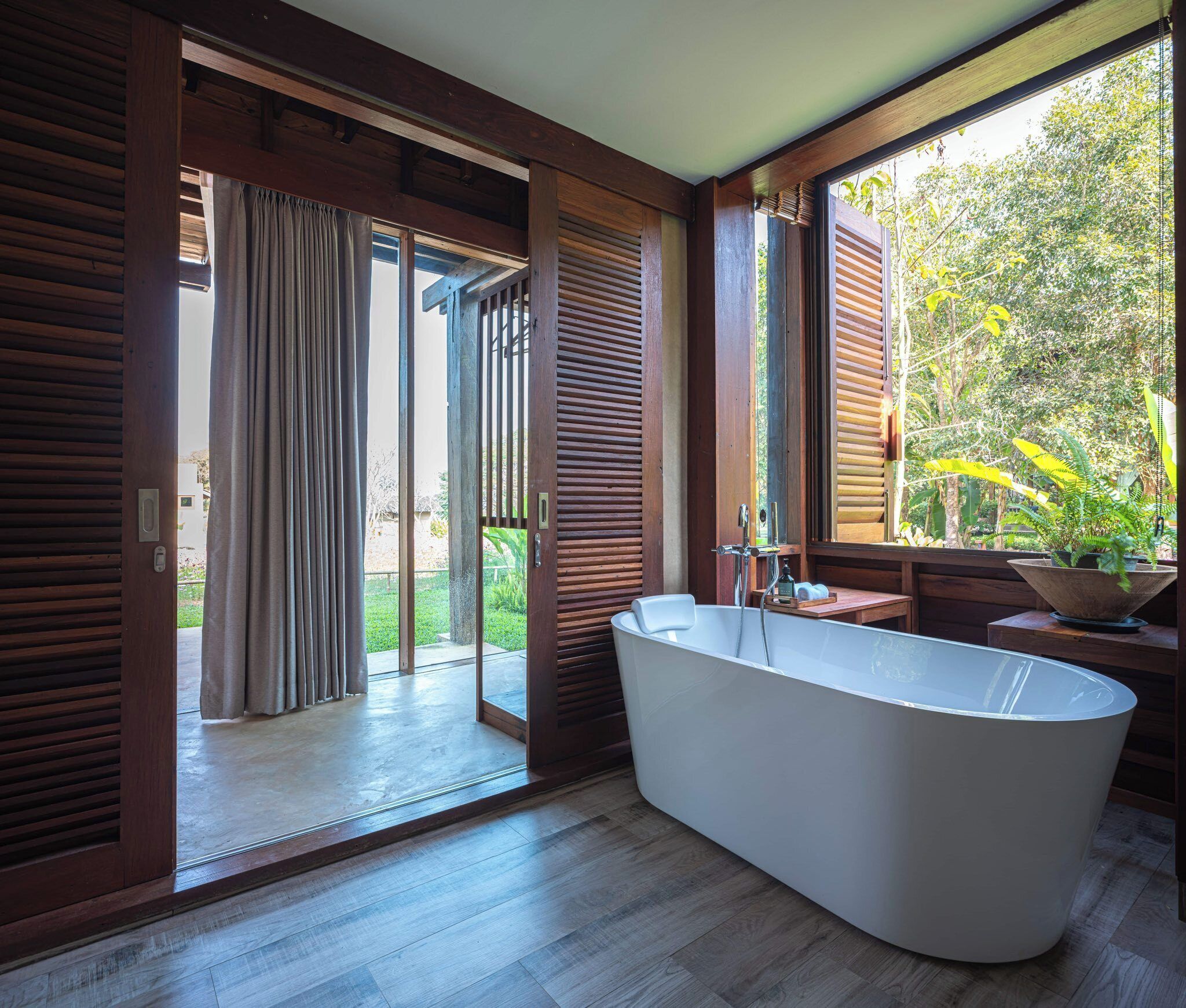 A modern bathroom with a freestanding bathtub, wooden flooring, and a large window offering a view of greenery outside.