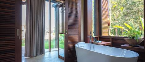 A modern bathroom with a freestanding bathtub, wooden flooring, and a large window offering a view of greenery outside.