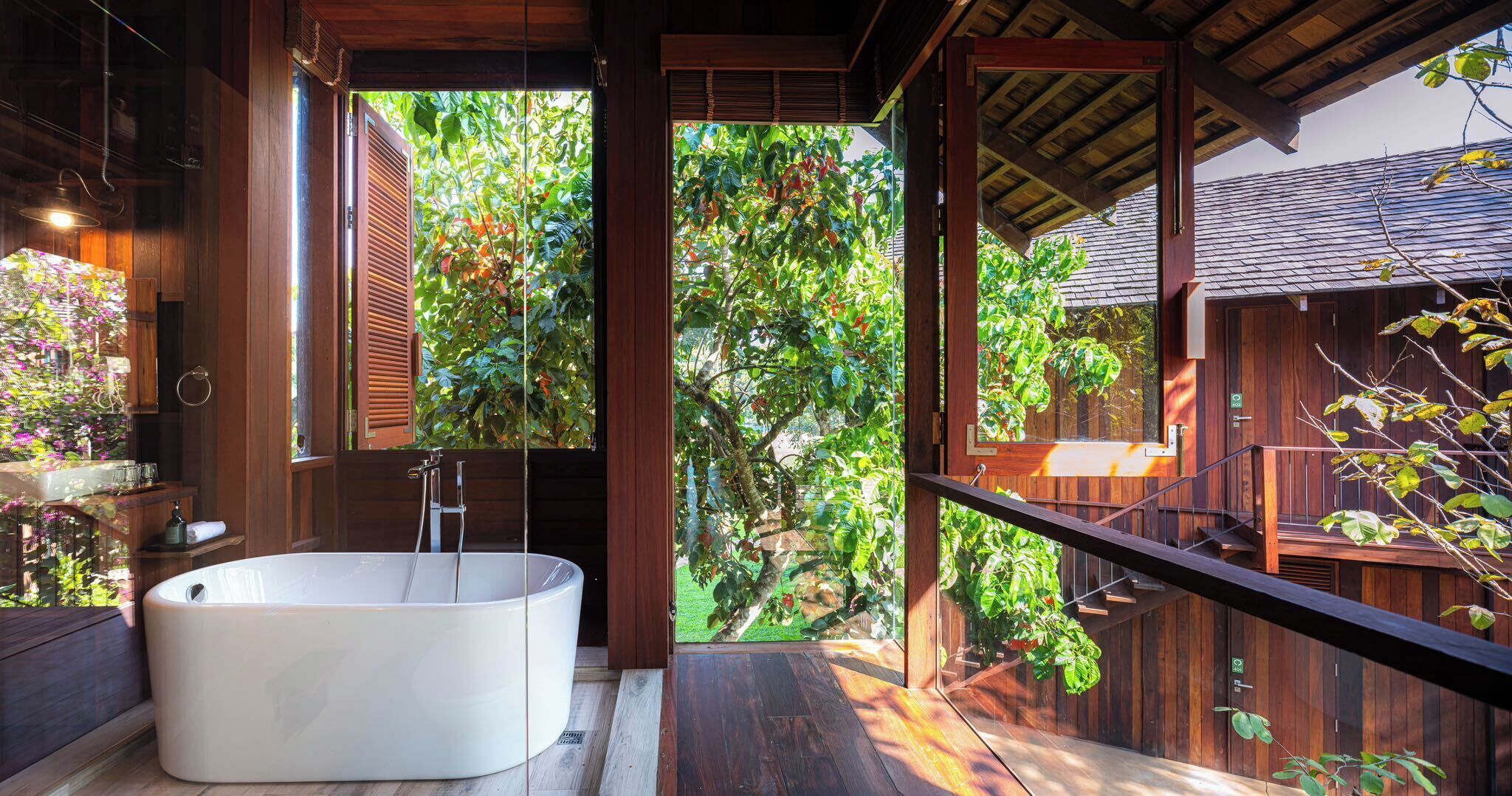 A modern bathroom with a freestanding bathtub, wooden flooring, and large windows offering views of lush greenery.