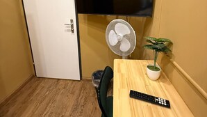 Ett litet rum med trägolv, en vit dörr, en tv som är monterad på väggen, ett bord med en fjärrkontroll, en stol, en fläkt och en krukväxt.