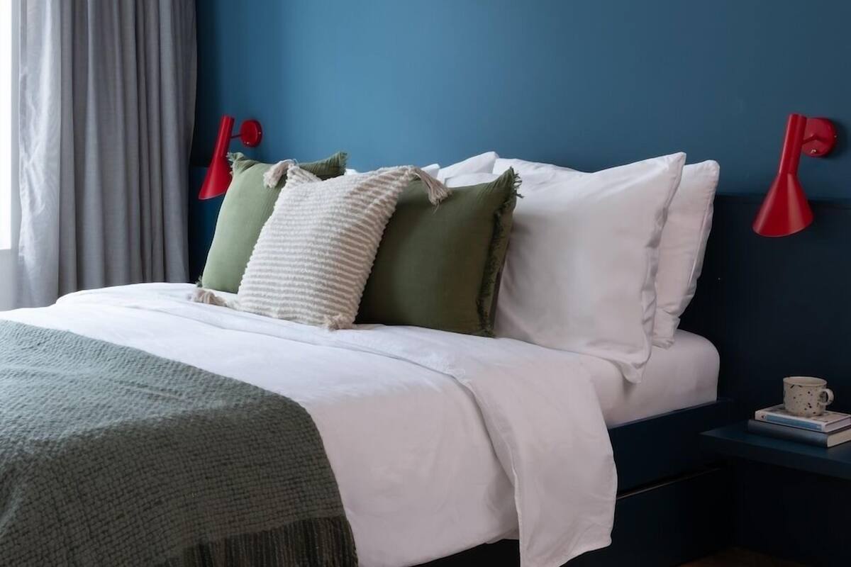 A bedroom with a bed, a nightstand, a red wall lamp, a ceiling light, and a window with curtains.