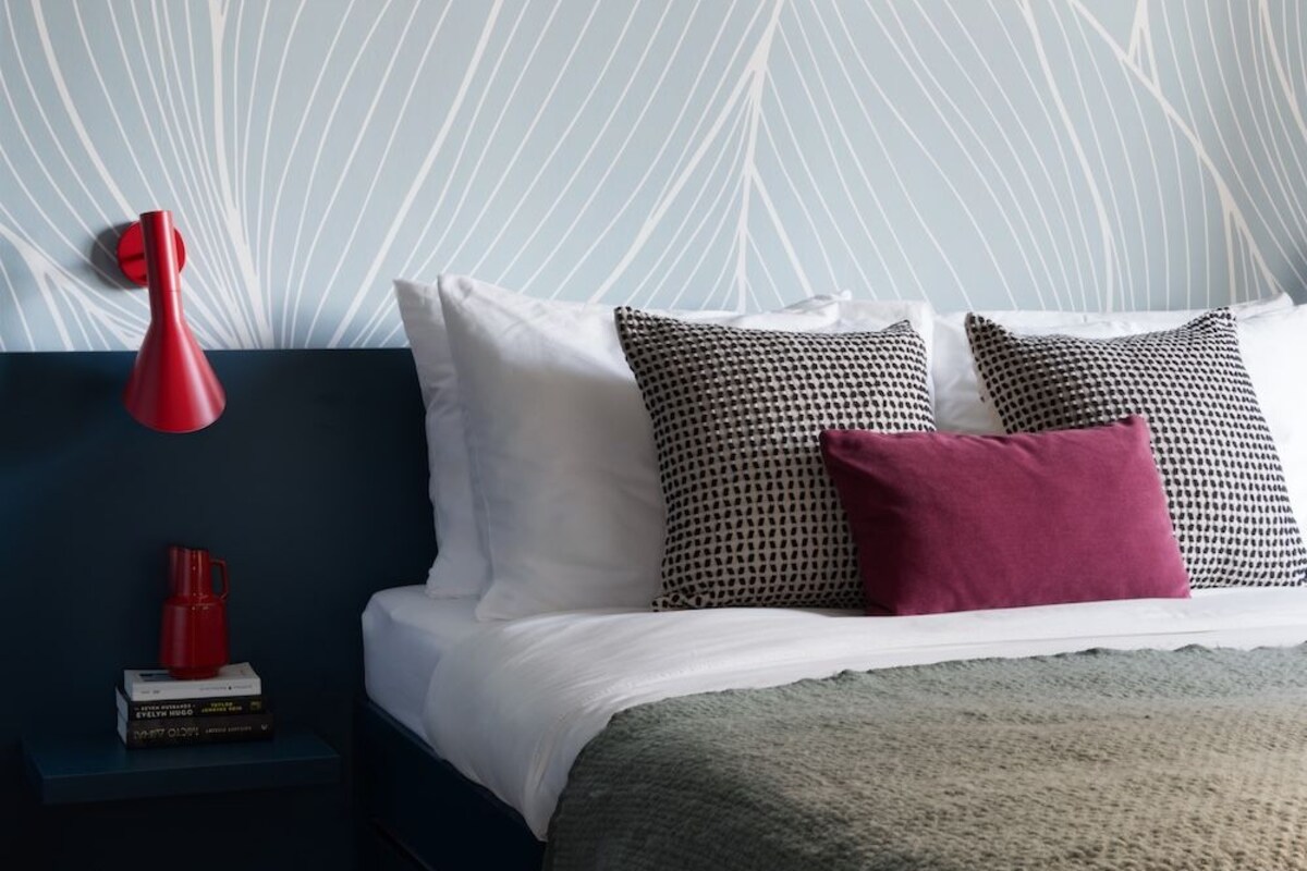 A bedroom with a bed, pillows, a bedside table with books, a red lamp, and a wall with a leaf pattern.