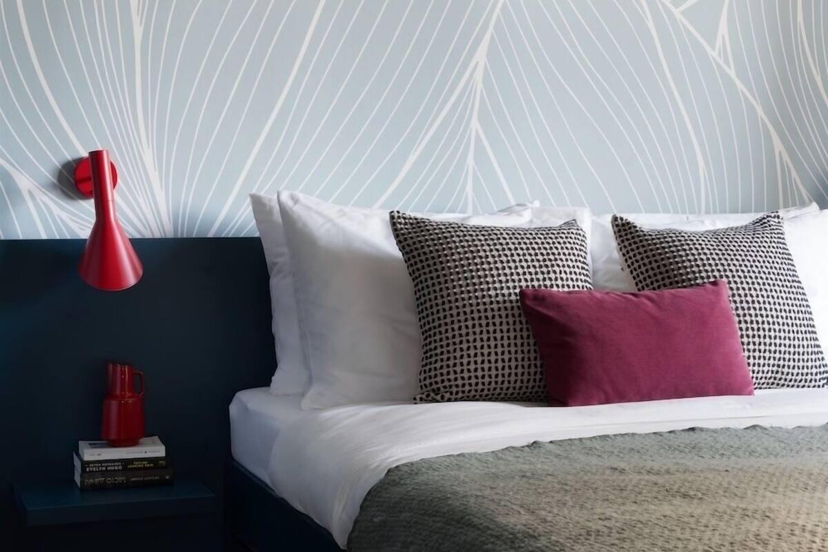 A bedroom with a bed, pillows, a bedside table with books, a red lamp, and a wall with a leaf pattern.