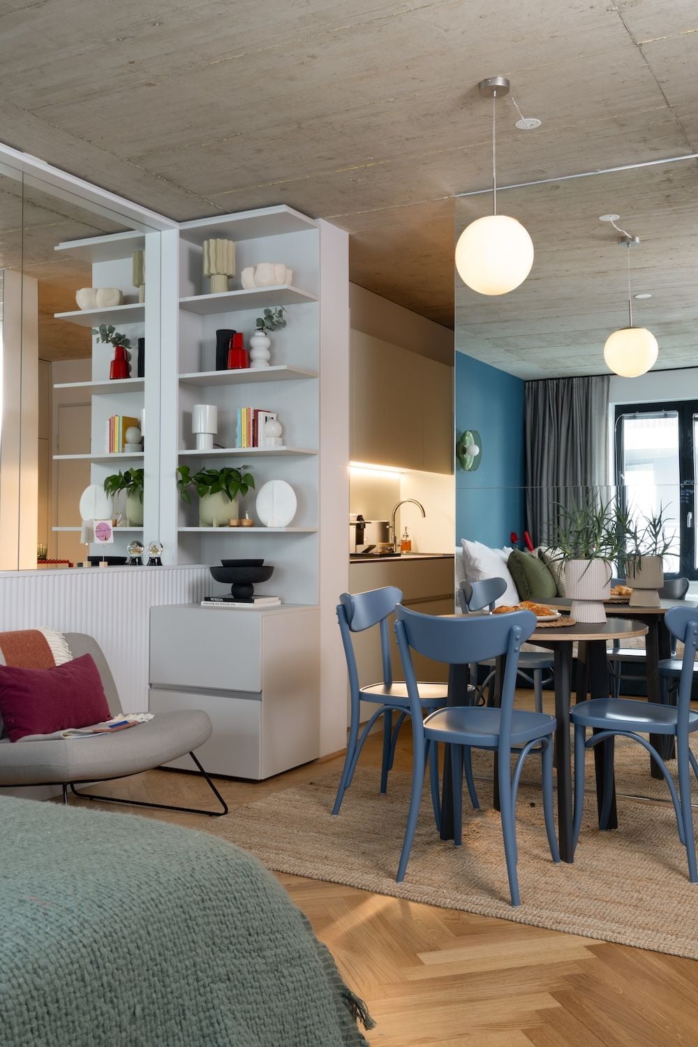 Ein modernes Wohnzimmer mit einer weißen Bücherwand, einem Essbereich mit blauen Stühlen und einem großen Fenster mit Vorhängen.