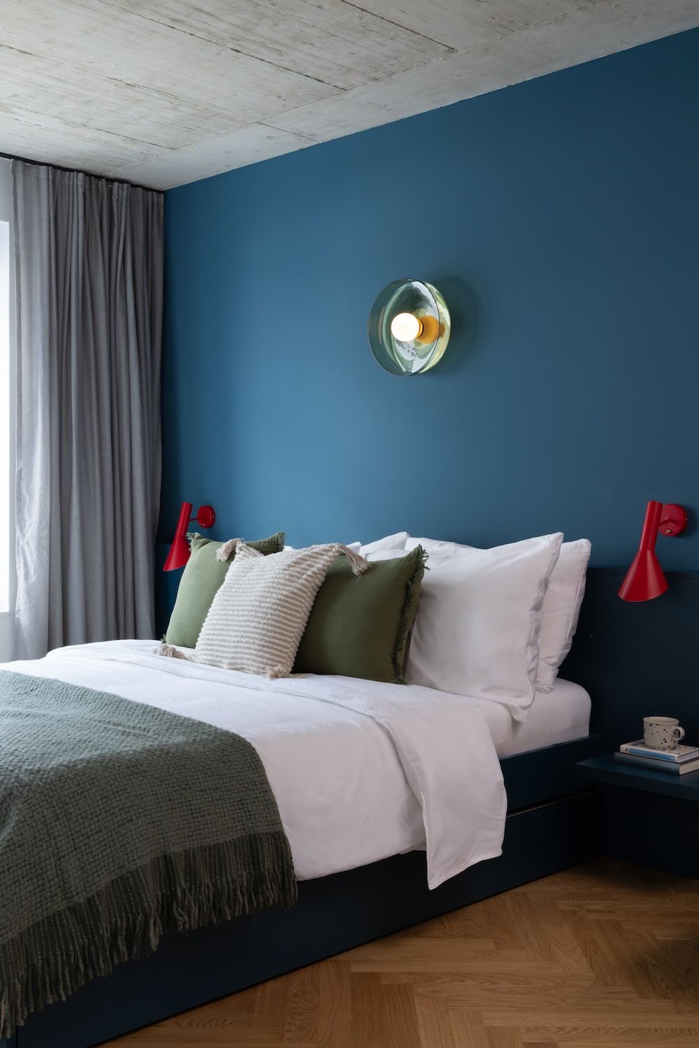 Een slaapkamer met een bed, een nachtkastje, een rode muurlamp, een plafondlamp en een raam met gordijnen.