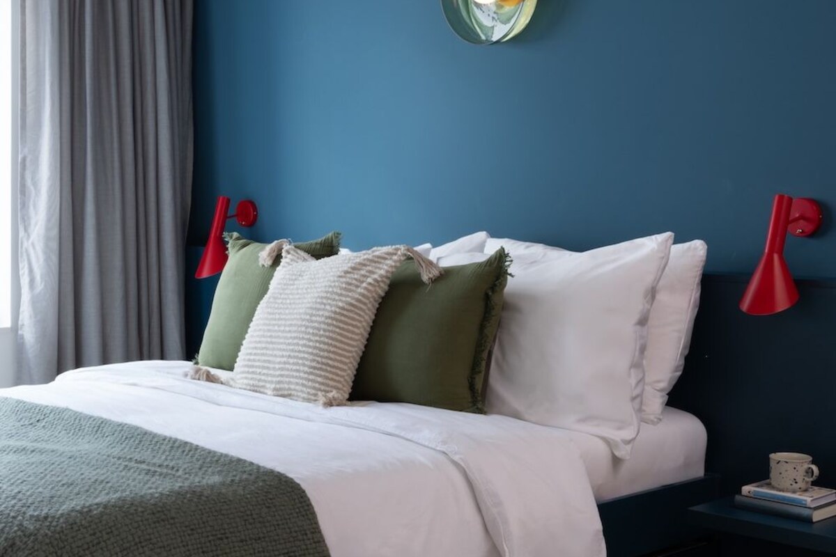A bedroom with a bed, a nightstand, a red wall lamp, a ceiling light, and a window with curtains.