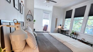 A bedroom with a bed, bedside table, lamp, and a large mirror. There is a window with blinds and a view of greenery outside. - Chic Algiers Point loft with king bed. Walk to ferry, minutes to French Quarter & jazz. (New Orleans)