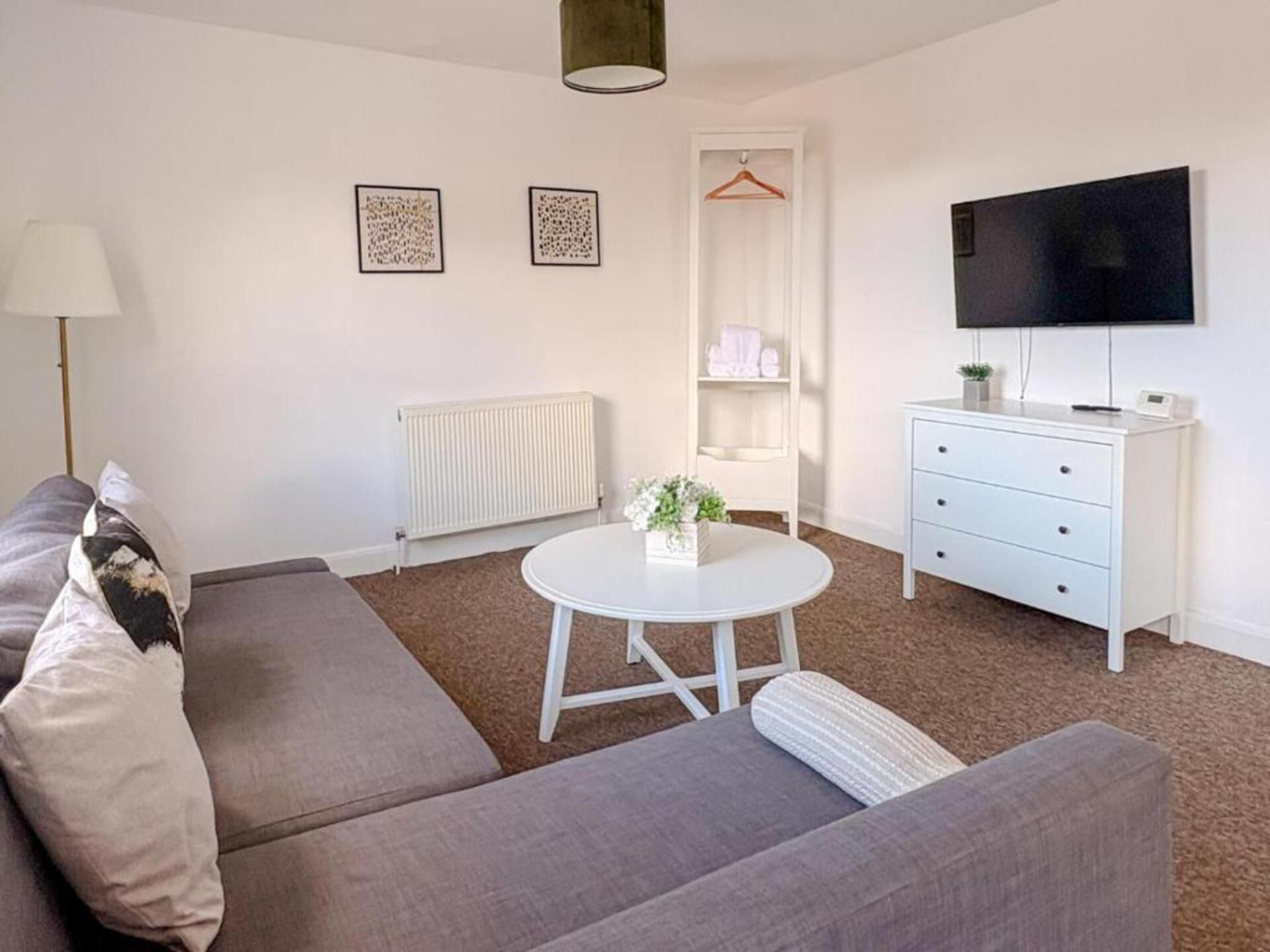 A modern living room with a grey sofa, a round white coffee table, a white dresser, a flat-screen TV, and two framed artworks on the wall.