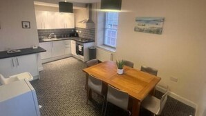 A compact kitchen with a dining area, featuring a tiled floor, a wooden dining table, and pendant lighting. - Church View Apartment - Nelson's Retreat (Great Yarmouth)