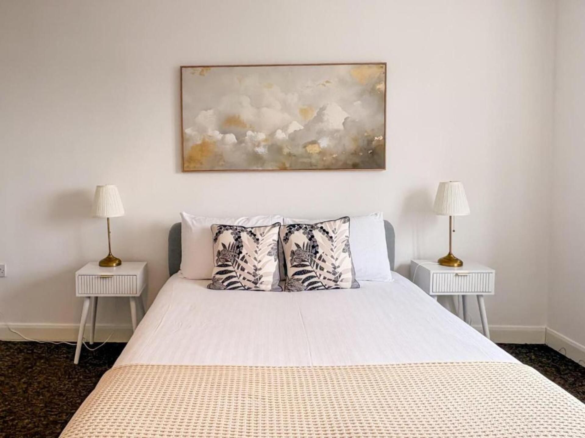 Habitación de hotel con una cama grande, dos mesitas de noche con lámparas, un cuadro abstracto colgado en la pared y una manta texturizada.