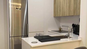 A compact kitchen with a stainless steel refrigerator, a white countertop, a wooden cabinet, a sink, and a bar stool.