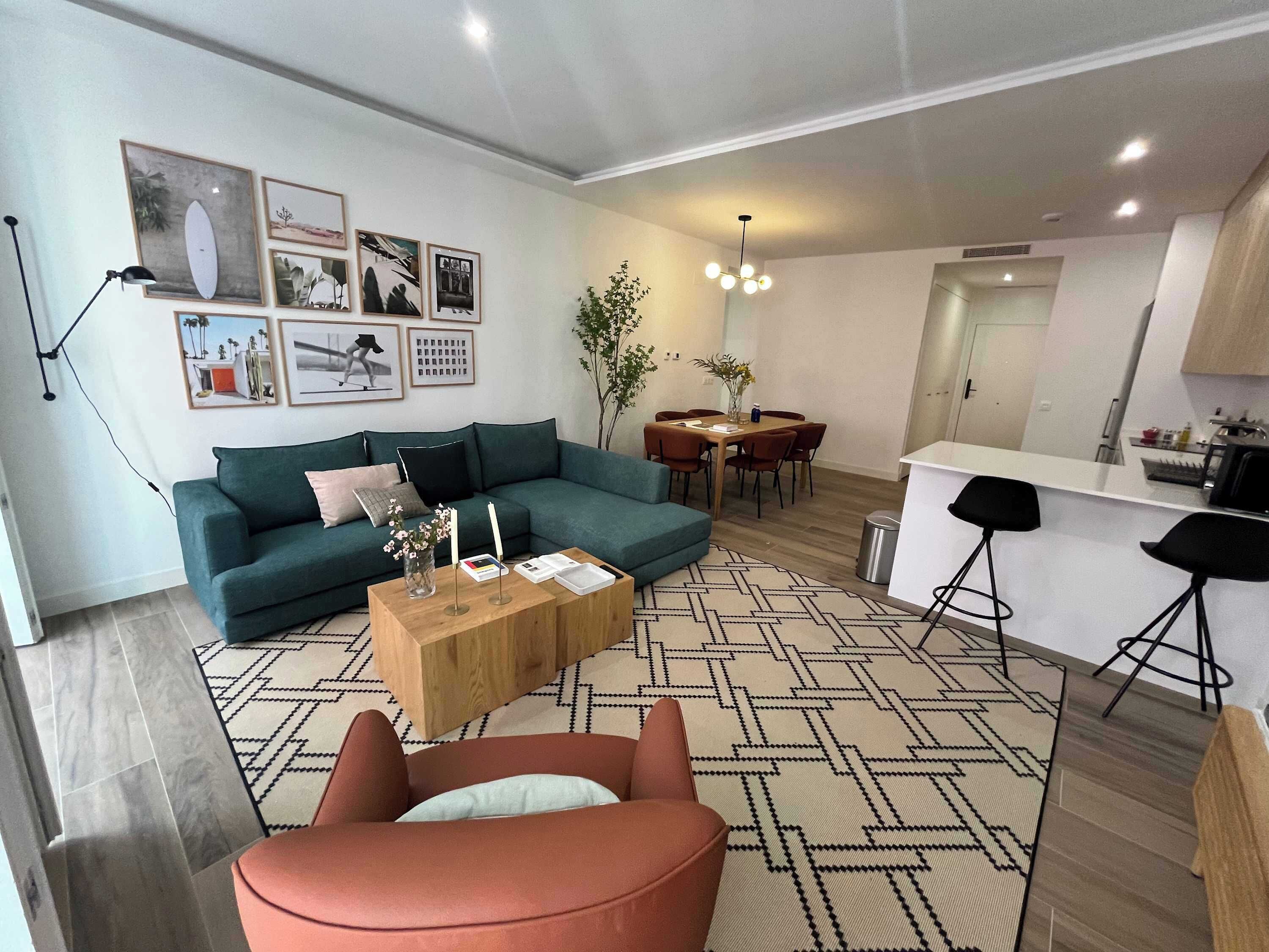 Una sala de estar moderna con un sofá verde, una mesa de centro y una silla roja. Hay un comedor con una mesa y sillas, y una cocina con una barra y taburetes.