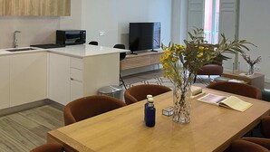 Et moderne køkken med hvide skabe, en mikroovn og et sort fjernsyn på et træskab. Der er et spisebord med stole og en vase med blomster.