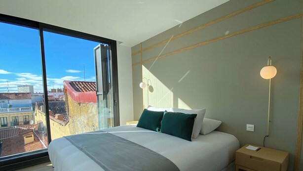Une chambre d’hôtel moderne, dotée d’un grand lit, d’une table de chevet, d’une lampe sur pied et offrant une vue sur la ville grâce à une grande fenêtre.