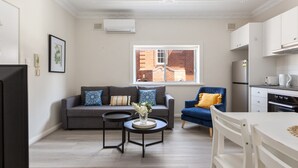 A modern living room with a grey sofa, a blue armchair, and a white dining table. There is a window with a view of a brick building. - Chic 1-bed Retreat Near Shops & Beach (Sydney)