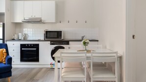A modern kitchen with white cabinetry, a built-in oven, microwave, and dishwasher. There is a dining table with chairs and a vase with flowers. - Chic 1-bed Retreat Near Shops & Beach (Sydney)