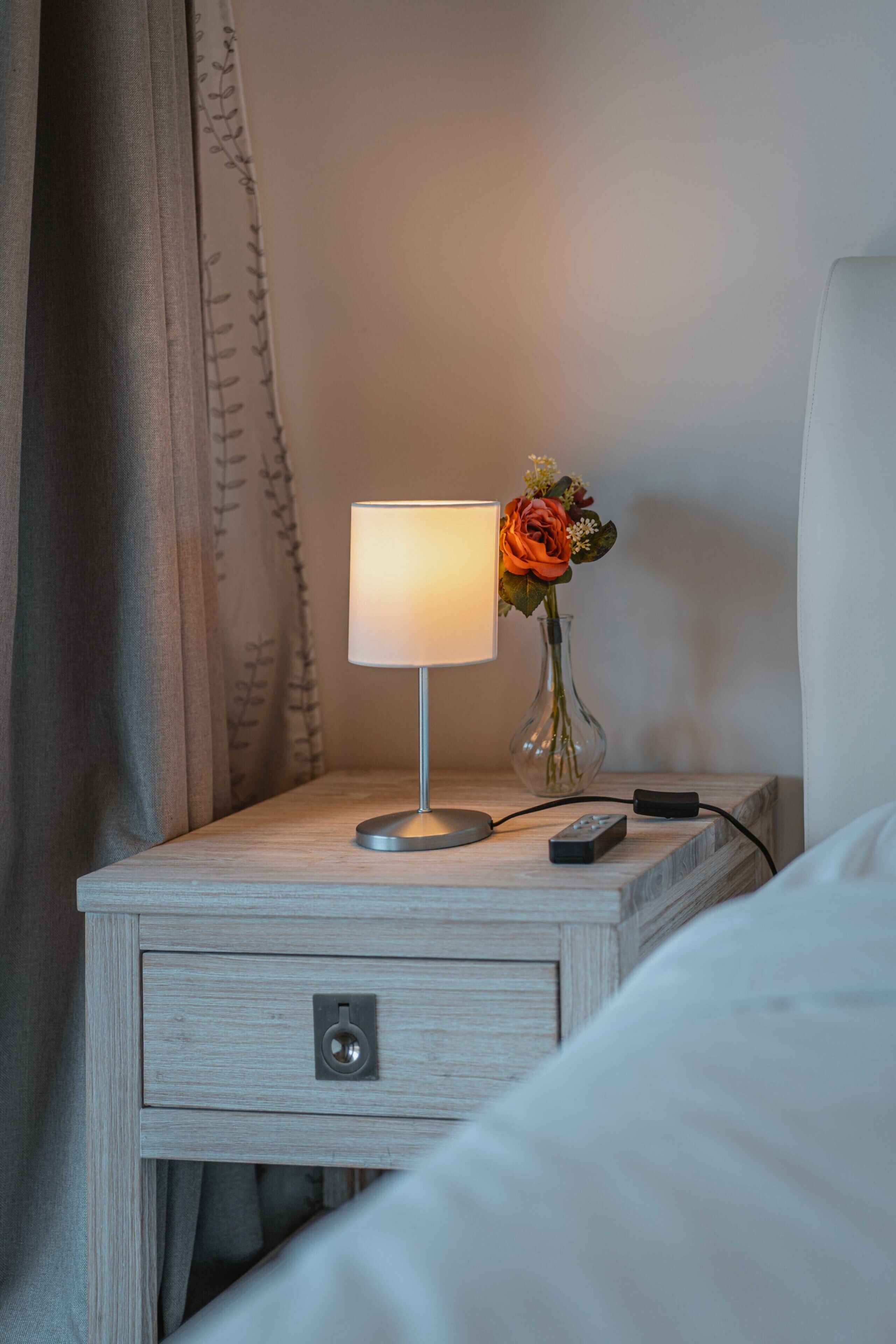 Een nachtkastje met een lamp, een vaas met bloemen en een afstandsbediening.