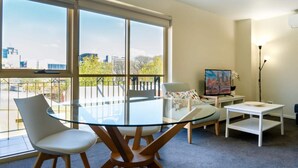 A modern living room with a glass dining table, two chairs, a small white side table, a television, and a painting on the wall. - Spacious City Stay Near Lygon Street Dining (Melbourne)