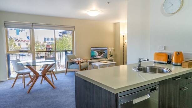 A modern kitchen with a built-in dishwasher, a sink, and a small dining area with a glass table and chairs. - Spacious City Stay Near Lygon Street Dining (Melbourne)