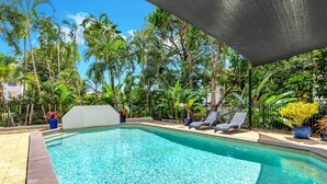 A rectangular swimming pool with clear blue water, surrounded by a paved deck and a black metal fence. There are lounge chairs under a shaded area, and various potted plants and trees are visible in the background. - Beachfront 3-bed With Pool & Stunning Ocean Views (Cairns)