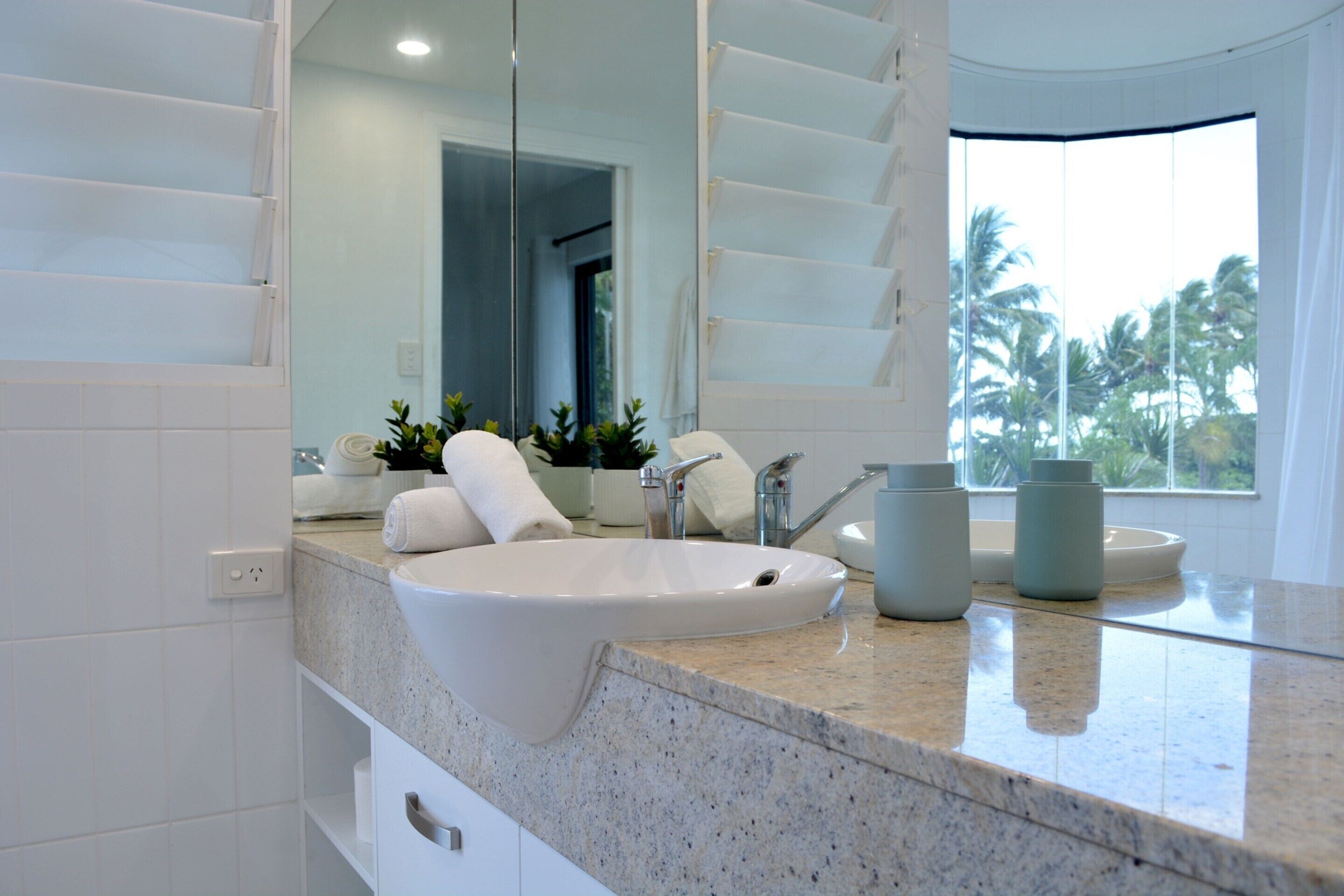 Une salle de bain moderne avec un grand lavabo blanc, un miroir et une fenêtre donnant sur des palmiers.