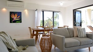 A modern living room with a sofa, a dining table with chairs, and a large window with curtains. - Beachfront 3-bed With Pool & Stunning Ocean Views (Cairns)