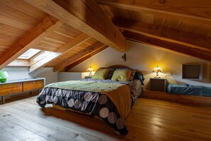 Une chambre avec un plafond en pente, des poutres en bois, un lit recouvert d’une couvre-lit à pois, deux lampes de chevet et un puits de lumière.
