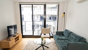A modern living room with a sofa, a glass coffee table, a TV on a wooden stand, and a balcony with chairs. - Idyllic 1-bed Near Popular Shopping Streets (Melbourne)