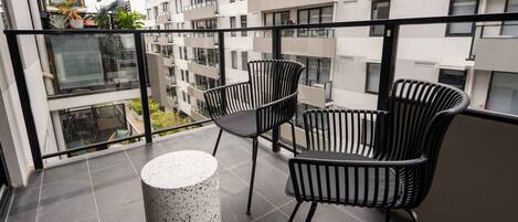 A balcony with two black chairs and a white round table, overlooking a cityscape with modern buildings.