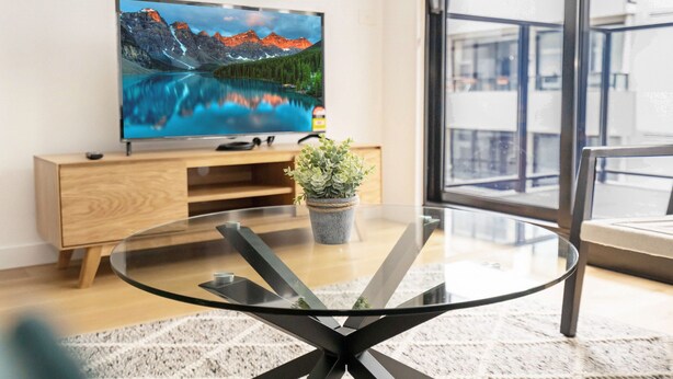 A modern living room with a flat-screen TV, a glass coffee table, a wooden cabinet, and a potted plant. - Idyllic 1-bed Near Popular Shopping Streets (Melbourne)