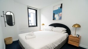 A neatly made bed with white linens, a bedside lamp, and a small wooden nightstand with an alarm clock. - Idyllic 1-bed Near Popular Shopping Streets (Melbourne)