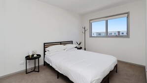 Une chambre avec un lit simple, une table de chevet, une lampe et une fenêtre donnant sur des immeubles.