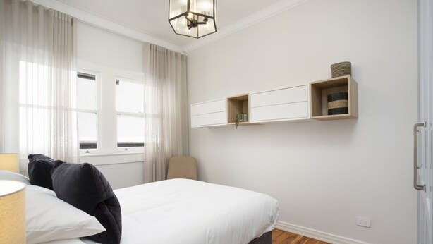 A bedroom with a bed, a chair, a bedside table, a lamp, a window with curtains, and built-in shelves with decorative items. - Coastal Living Just Metres From Coogee Beach (Sydney)