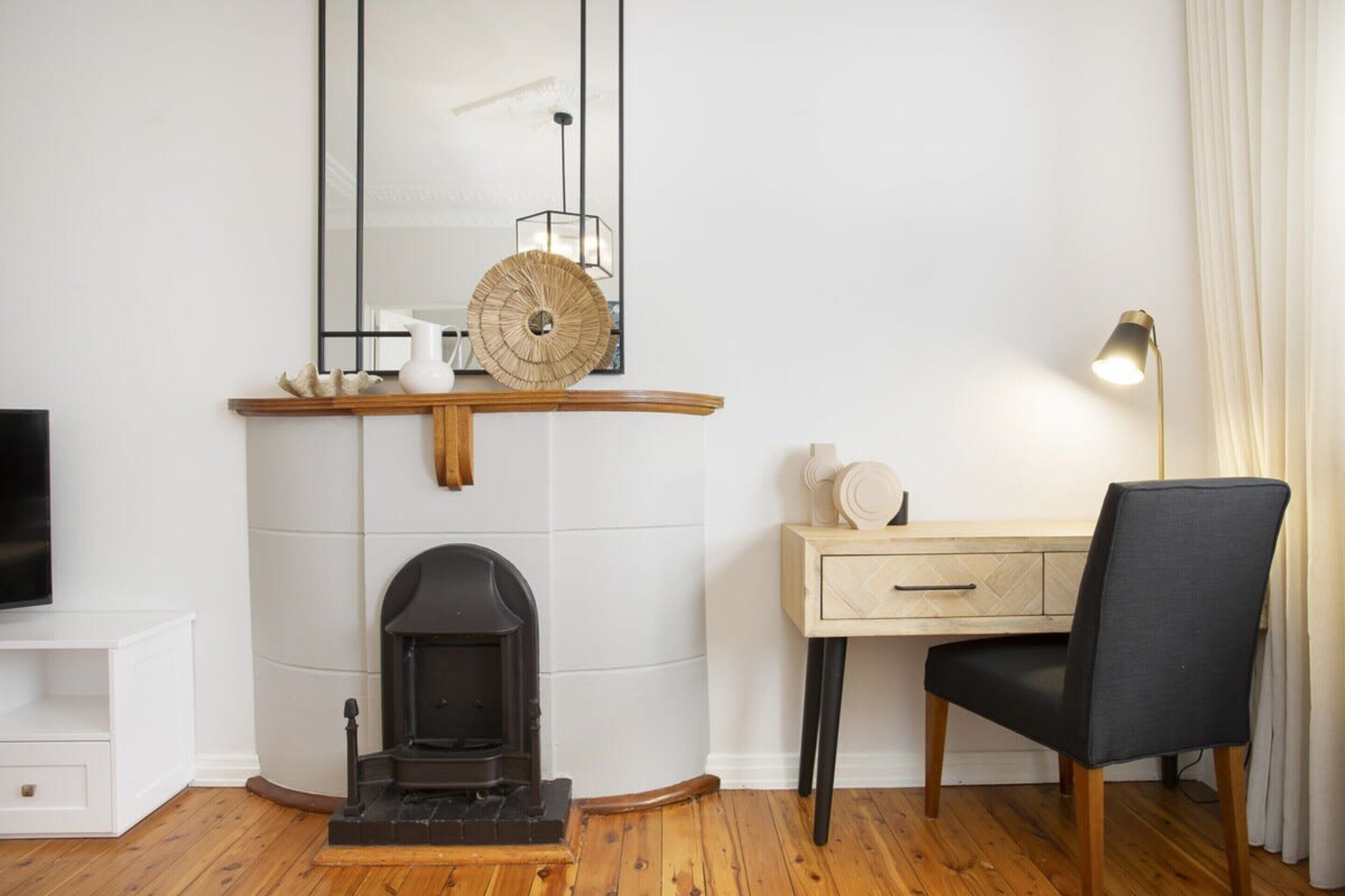 Un salon moderne avec une cheminée, une étagère en bois ornée d’objets décoratifs, un bureau avec une chaise et un téléviseur posé sur un meuble blanc.