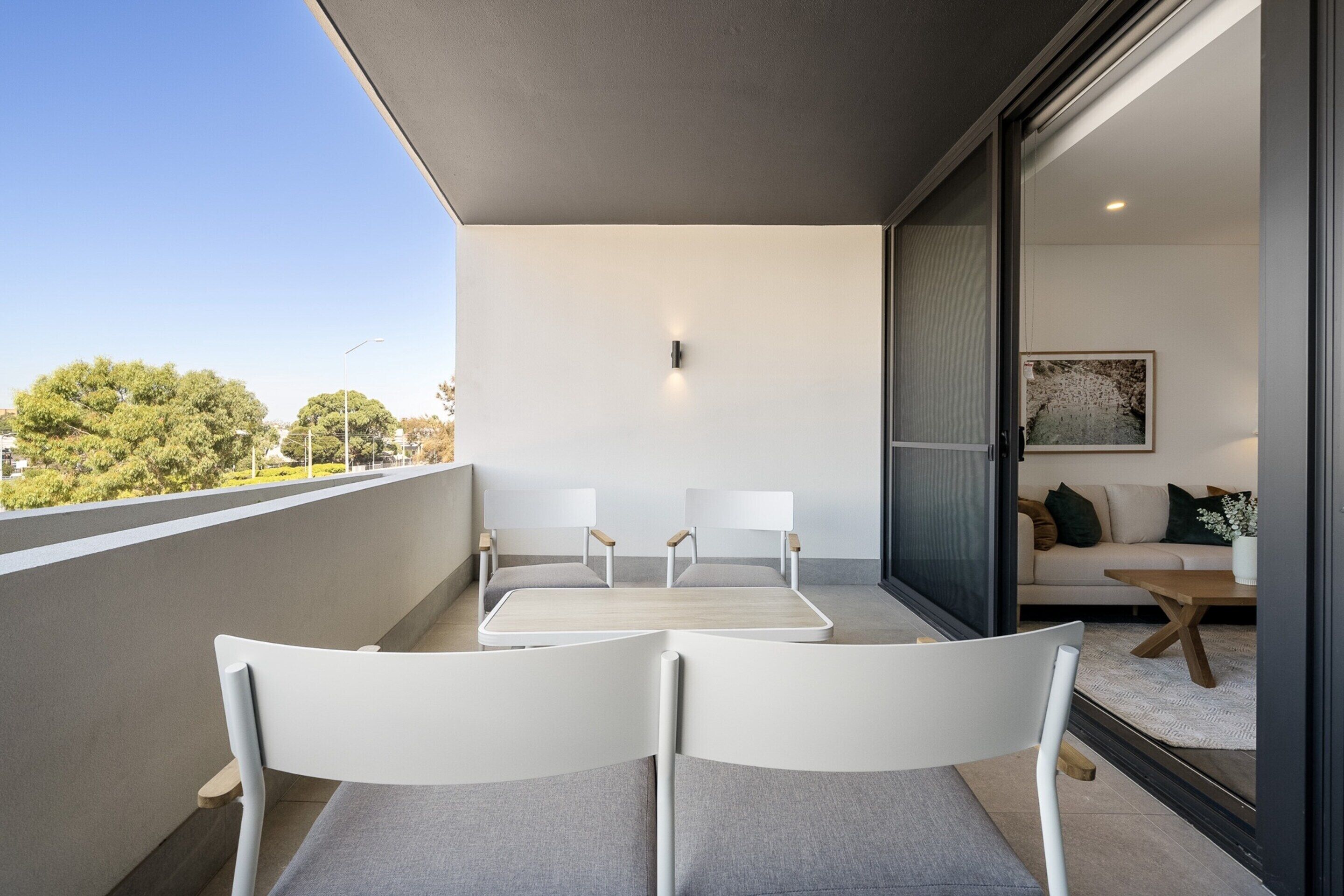 A modern balcony with two white chairs and a small table, a sofa with cushions, and a painting on the wall.
