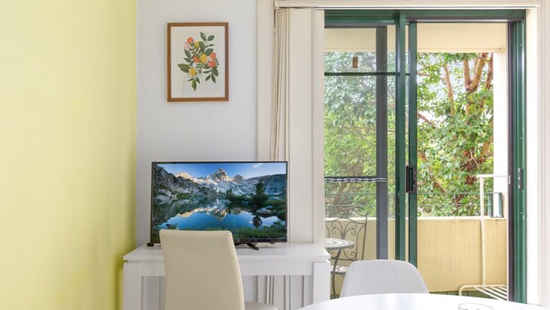 A room with a TV on a white stand, a dining table with chairs, and a balcony with a view of trees. - Serene Studio With Balcony in Prime Location (Sydney)