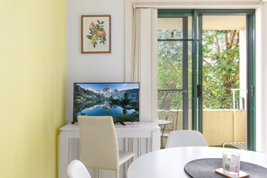 A room with a TV on a white stand, a dining table with chairs, and a balcony with a view of trees. - Serene Studio With Balcony in Prime Location (Sydney)