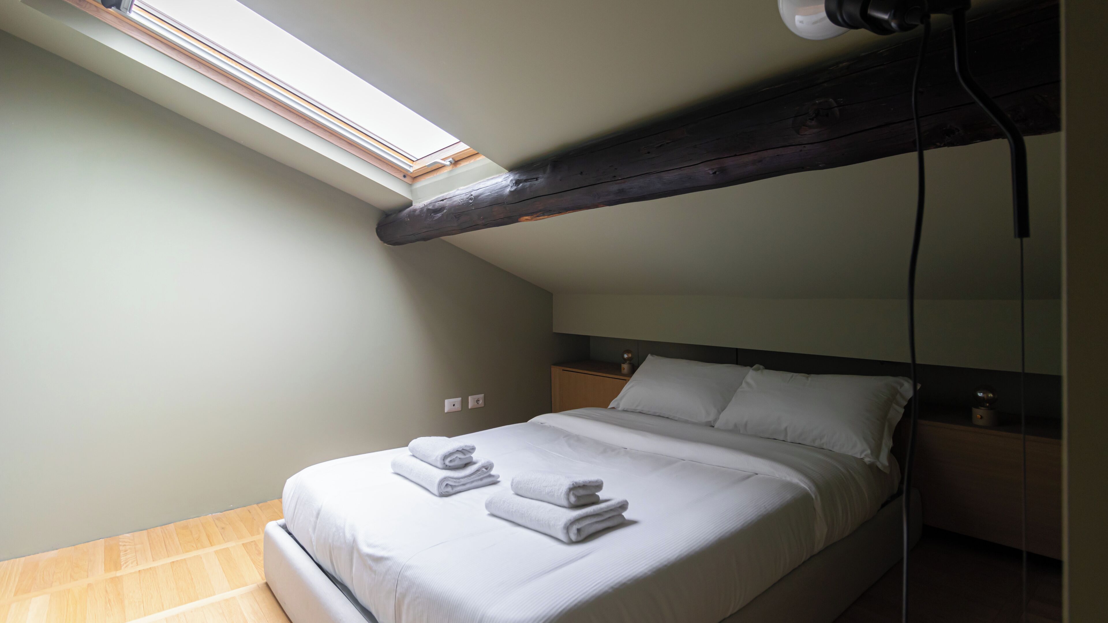 Une chambre avec un puits de lumière, un lit recouvert de draps blancs et un plancher en bois.